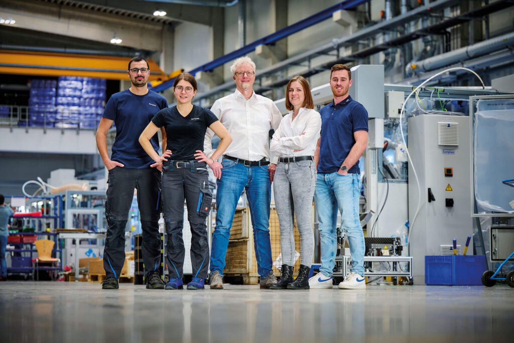 Es ist eine Gruppe von fünf Menschen, die in der Montagehalle von Sonplas stehen, zu sehen. Dabei handelt es sich um zwei Frauen und drei Männer. Jeweils eine Frau und und zwei Männer tragen Büro-Kleidung, sie sind Teil des Konstruktionsteams. Die restlichen Personen gehören zur Montageabteilung und tragen klassische Arbeitskleidung.