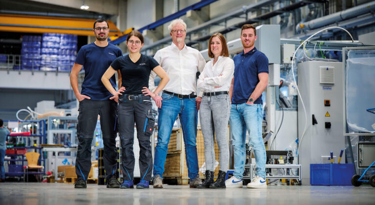 Es ist eine Gruppe von fünf Menschen, die in der Montagehalle von Sonplas stehen, zu sehen. Dabei handelt es sich um zwei Frauen und drei Männer. Jeweils eine Frau und und zwei Männer tragen Büro-Kleidung, sie sind Teil des Konstruktionsteams. Die restlichen Personen gehören zur Montageabteilung und tragen klassische Arbeitskleidung.