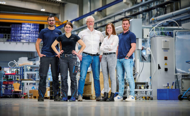 Es ist eine Gruppe von fünf Menschen, die in der Montagehalle von Sonplas stehen, zu sehen. Dabei handelt es sich um zwei Frauen und drei Männer. Jeweils eine Frau und und zwei Männer tragen Büro-Kleidung, sie sind Teil des Konstruktionsteams. Die restlichen Personen gehören zur Montageabteilung und tragen klassische Arbeitskleidung.