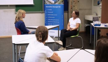Zu sehen ist Bettina Liegl und eine Schülerin des Anton-Bruckner-Gymnasiums. Sie sitzen gegenüber an einem Tisch und üben ein Bewerbergespräch.