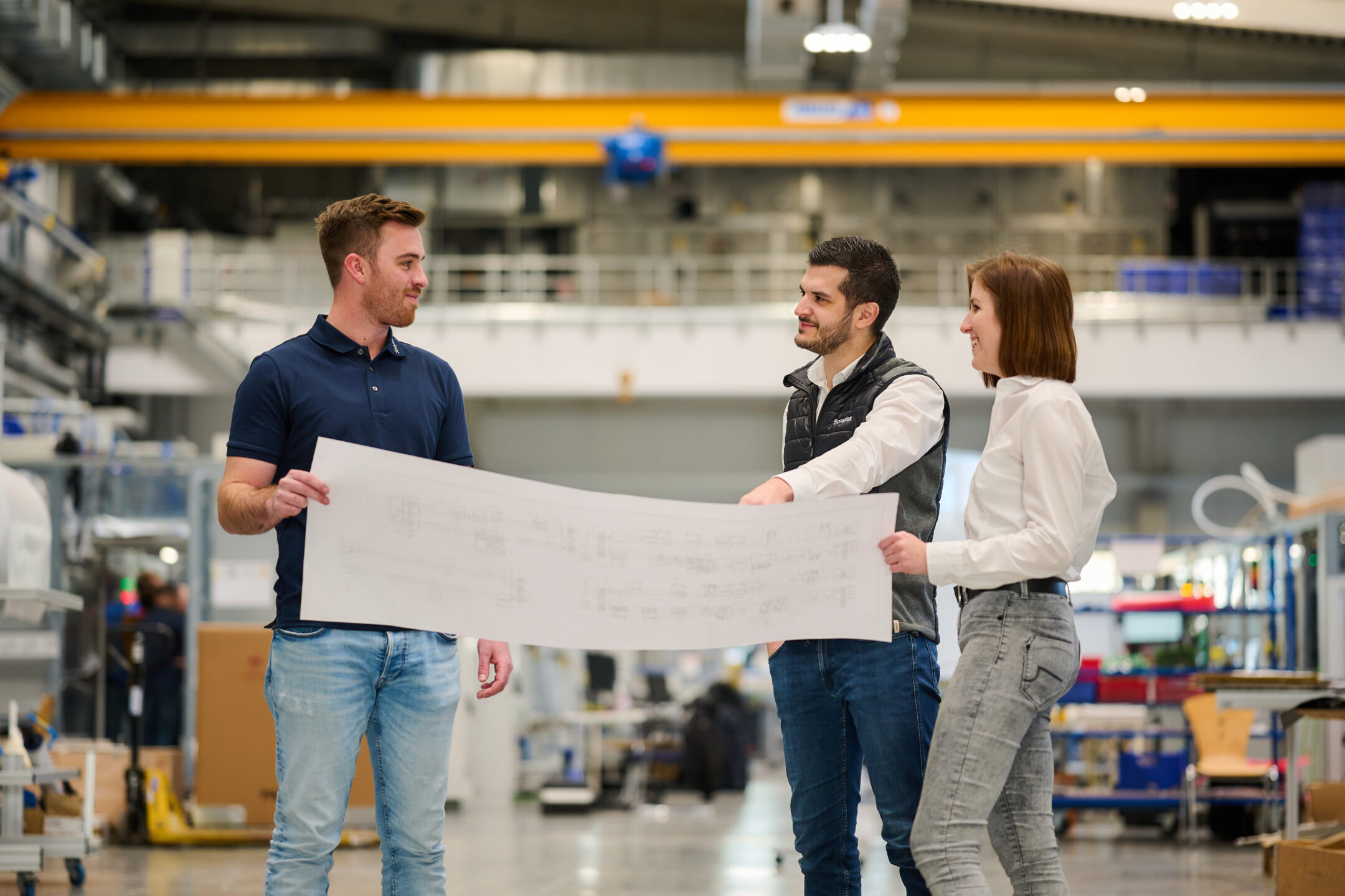 Drei Personen, darunter zwei Männer und eine Frau, stehen in der Montagehalle, halten einen Konstruktionsplan und schauen sich gegenseitig an